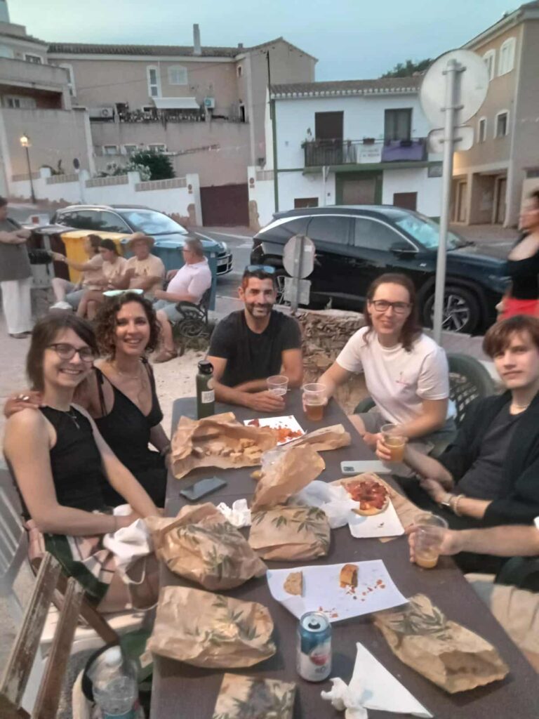 Group of people sitting around a table sharing food and drinks at the outdoor market, with cars and village buildings in the background