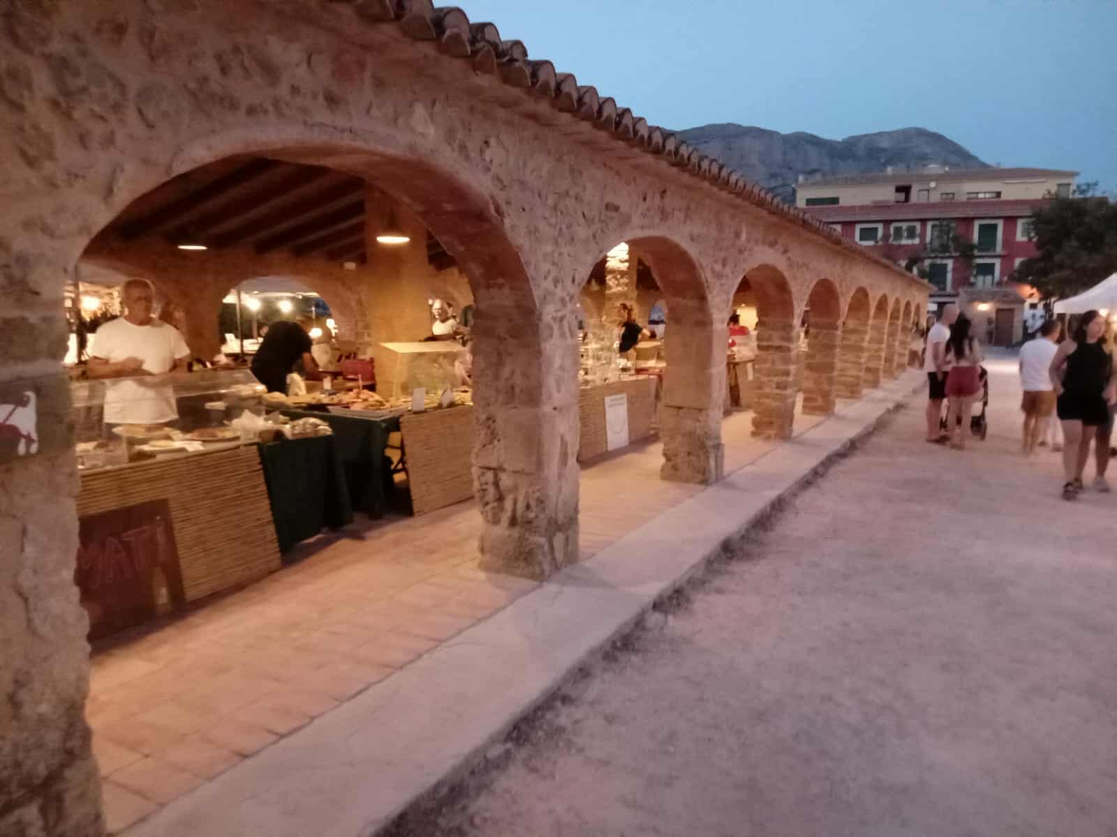 People strolling through the illuminated Riurau market in the evening, with warm lighting highlighting the stone archways