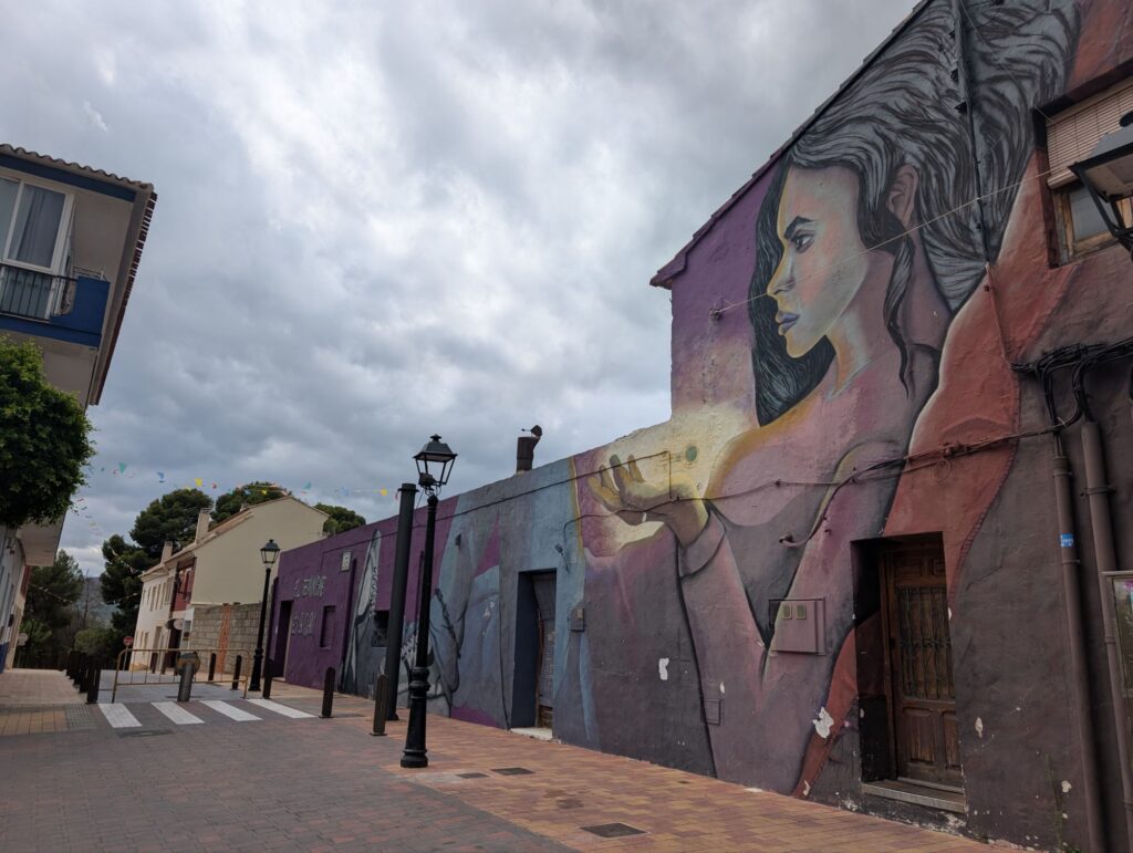 Large mural of a woman's portrait painted on white village buildings, with traditional Spanish street lamps and narrow cobbled streets